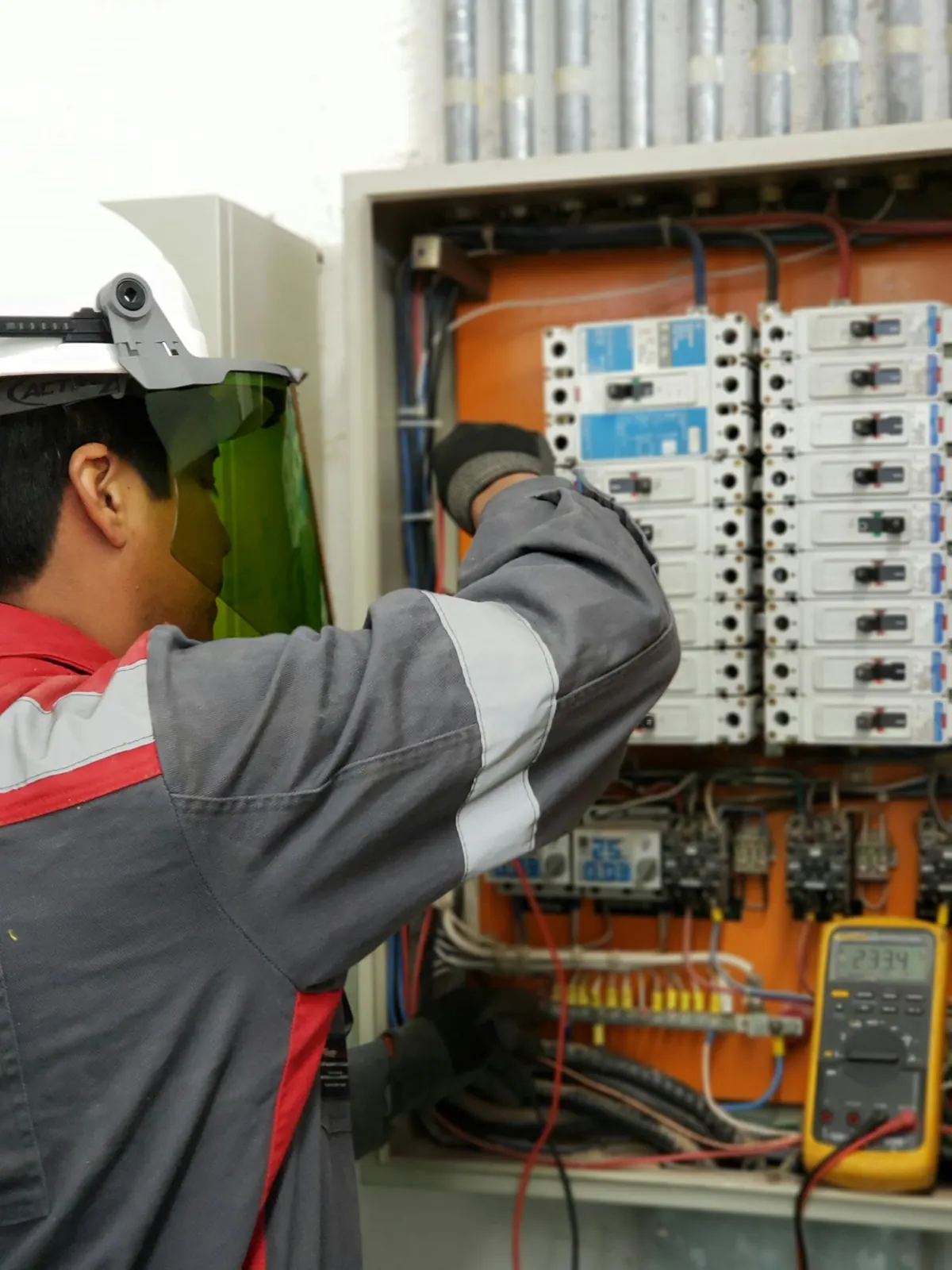 Eletricista com capacete e protetor facial medindo tensão em painel elétrico energizado